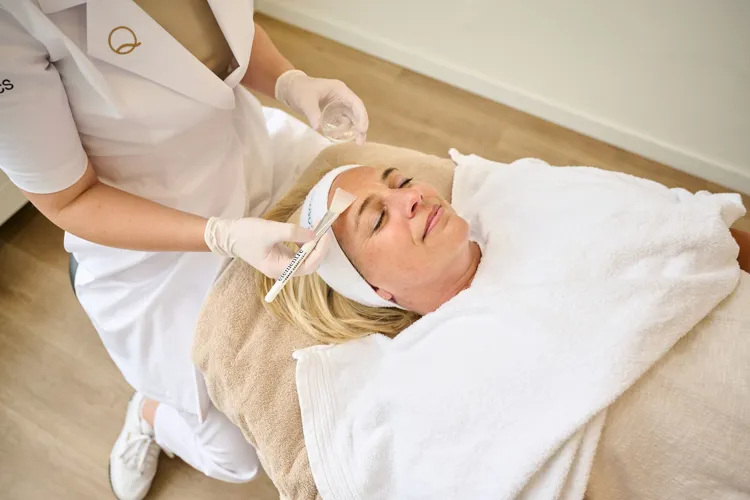 Frau entspannt sich mit geschlossenen Augen, während eine Kosmetikerin ein Produkt mit einem Pinsel auf ihre Stirn aufträgt.