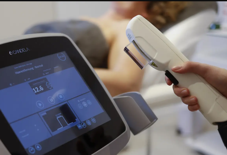 Nahaufnahme eines IPL-Geräts zur Haarentfernung mit sichtbarem Touchscreen und Handstück, das von einer Person gehalten wird, während im Hintergrund eine Person auf einer Behandlungsliege liegt.