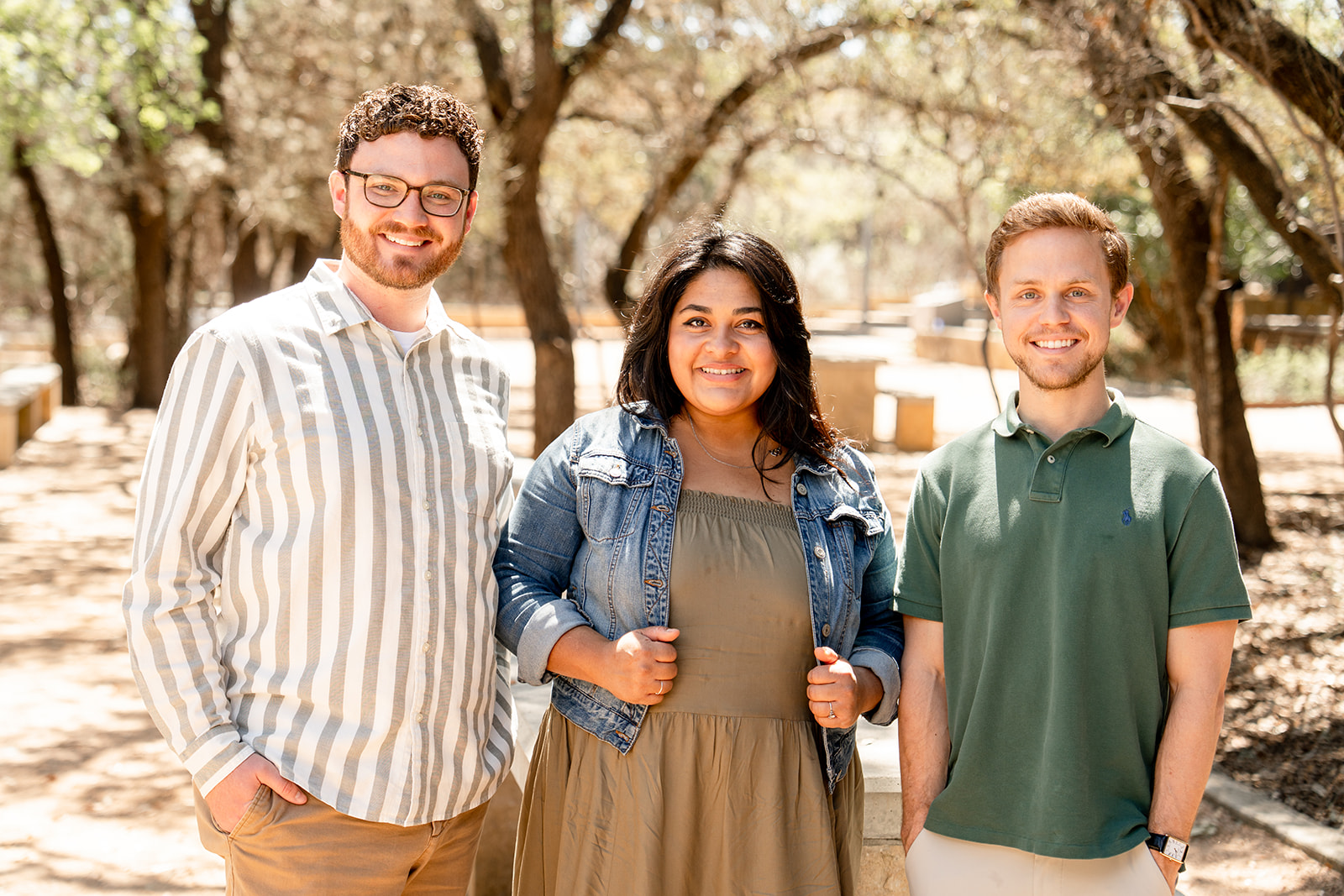Three smiling young adults standing outdoors among trees, two men and one woman in casual clothes.