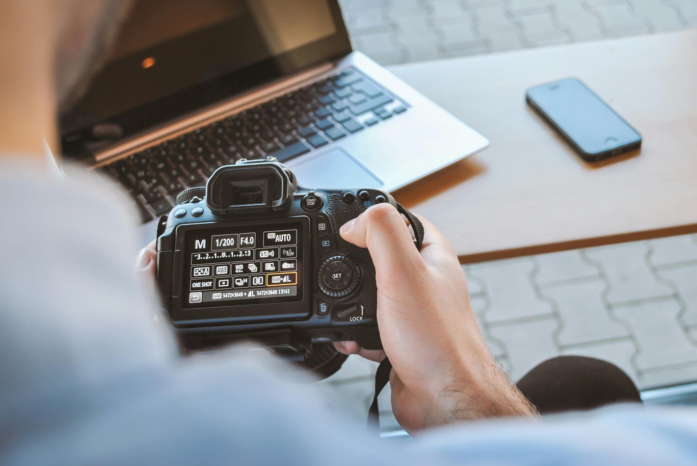 An over-the-shoulder look at a man browsing his camera's settings while sitting at a table with his laptop.