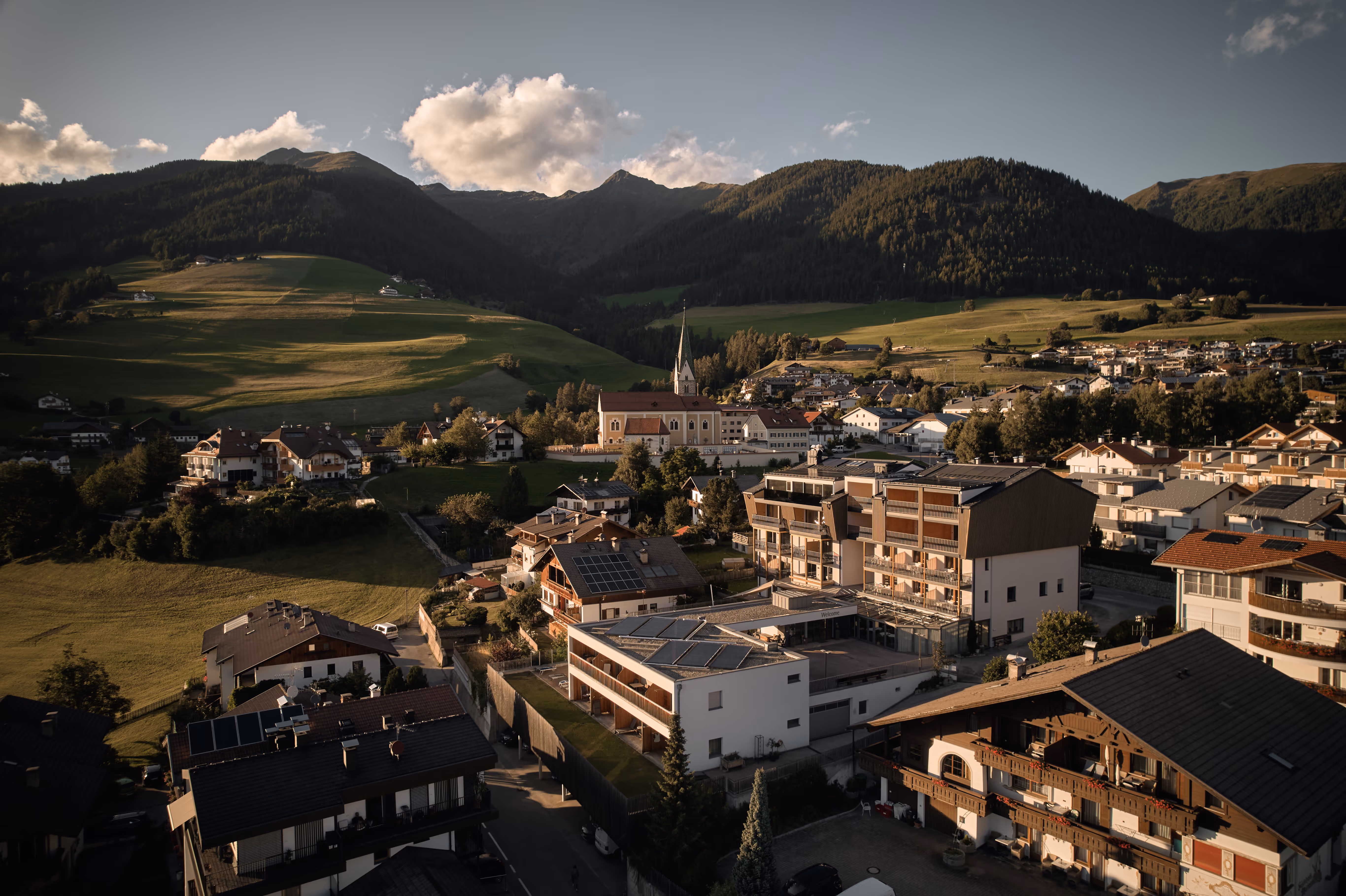 Marketing- und Social-Media-Projekt Montara: Blick auf ein Dorf inmitten grüner Hügel und Berge bei tiefstehender Abendsonne, mit Kirche im Zentrum.