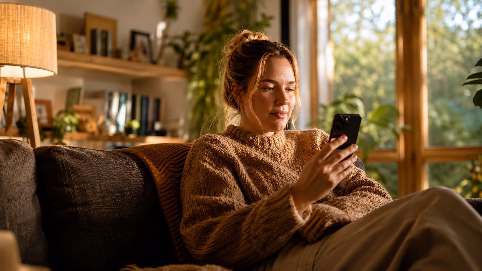 Woman in a brown sweater sitting on a couch looking at a smartphone in a cozy room with plants.