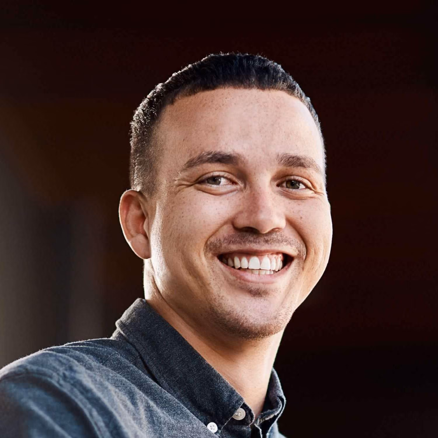 Smiling man with short dark hair and a buttoned gray shirt against a dark background.