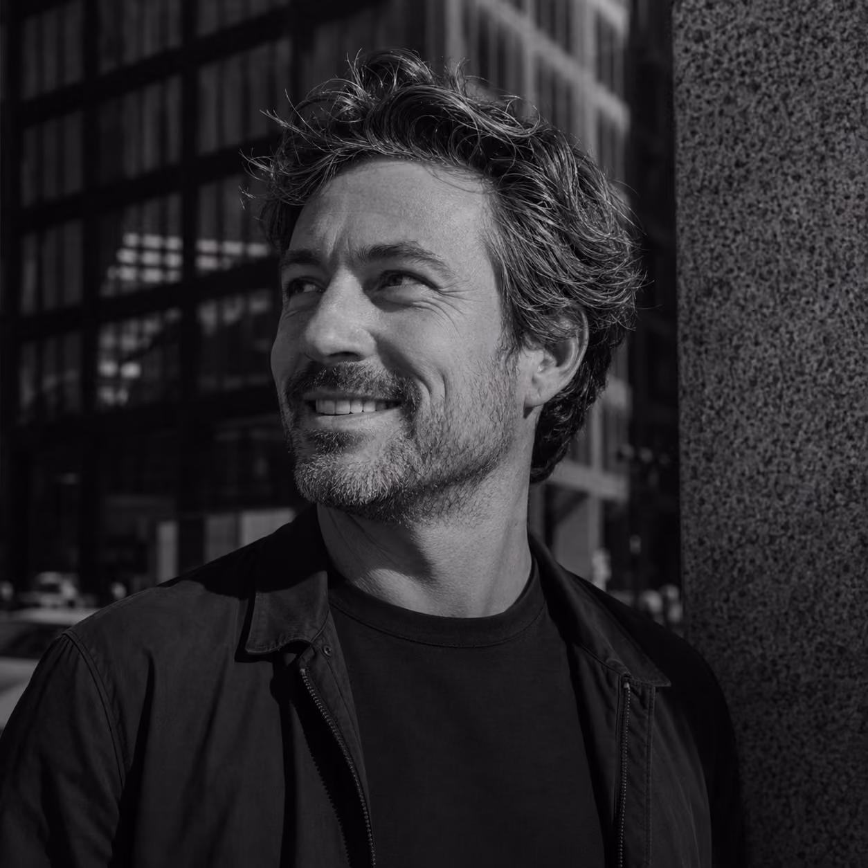 Smiling man with wavy hair and beard standing outdoors by a textured wall in a city setting.