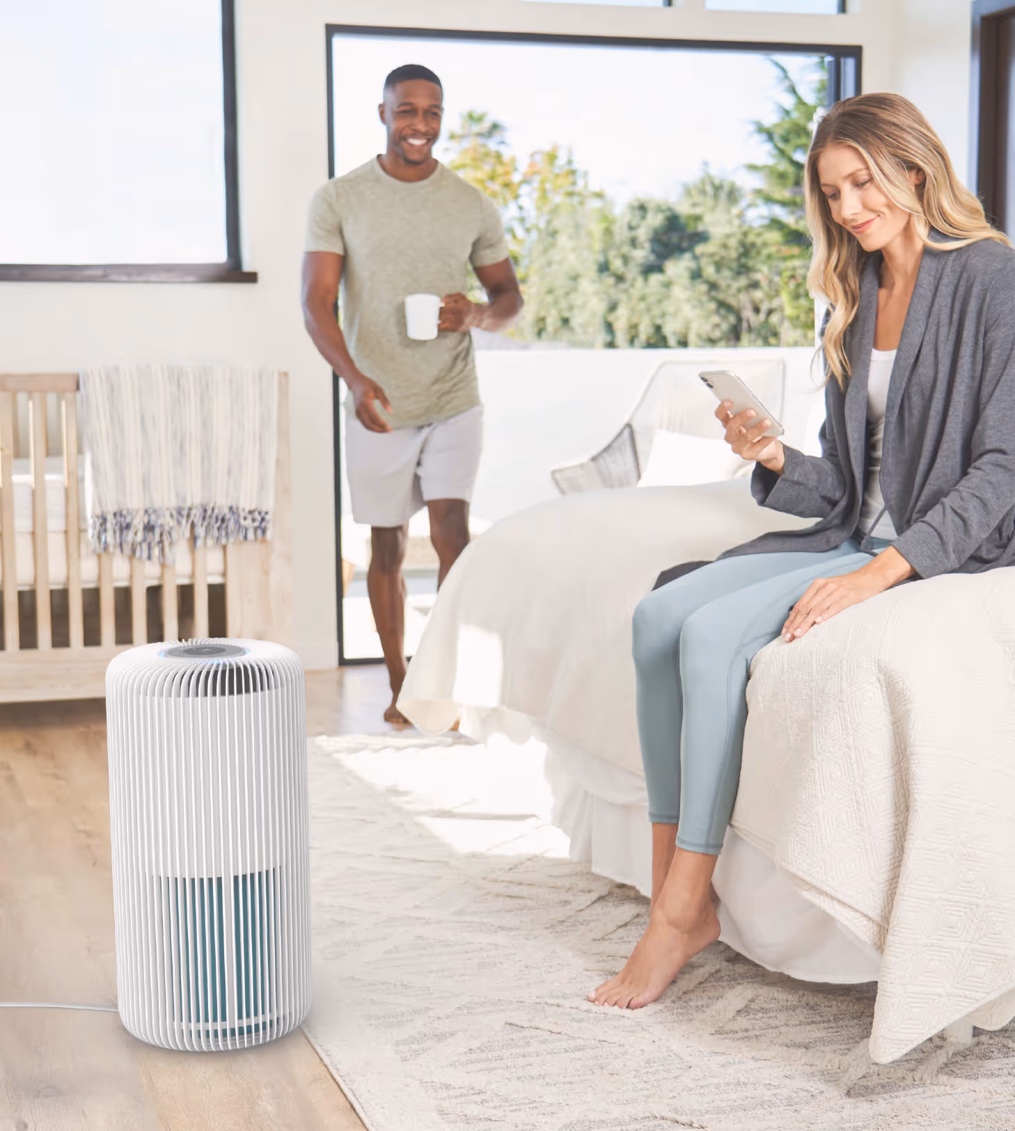 Modern white air purifier on the floor in a bright bedroom with a woman sitting on the bed looking at her phone and a man holding a cup walking in the background.