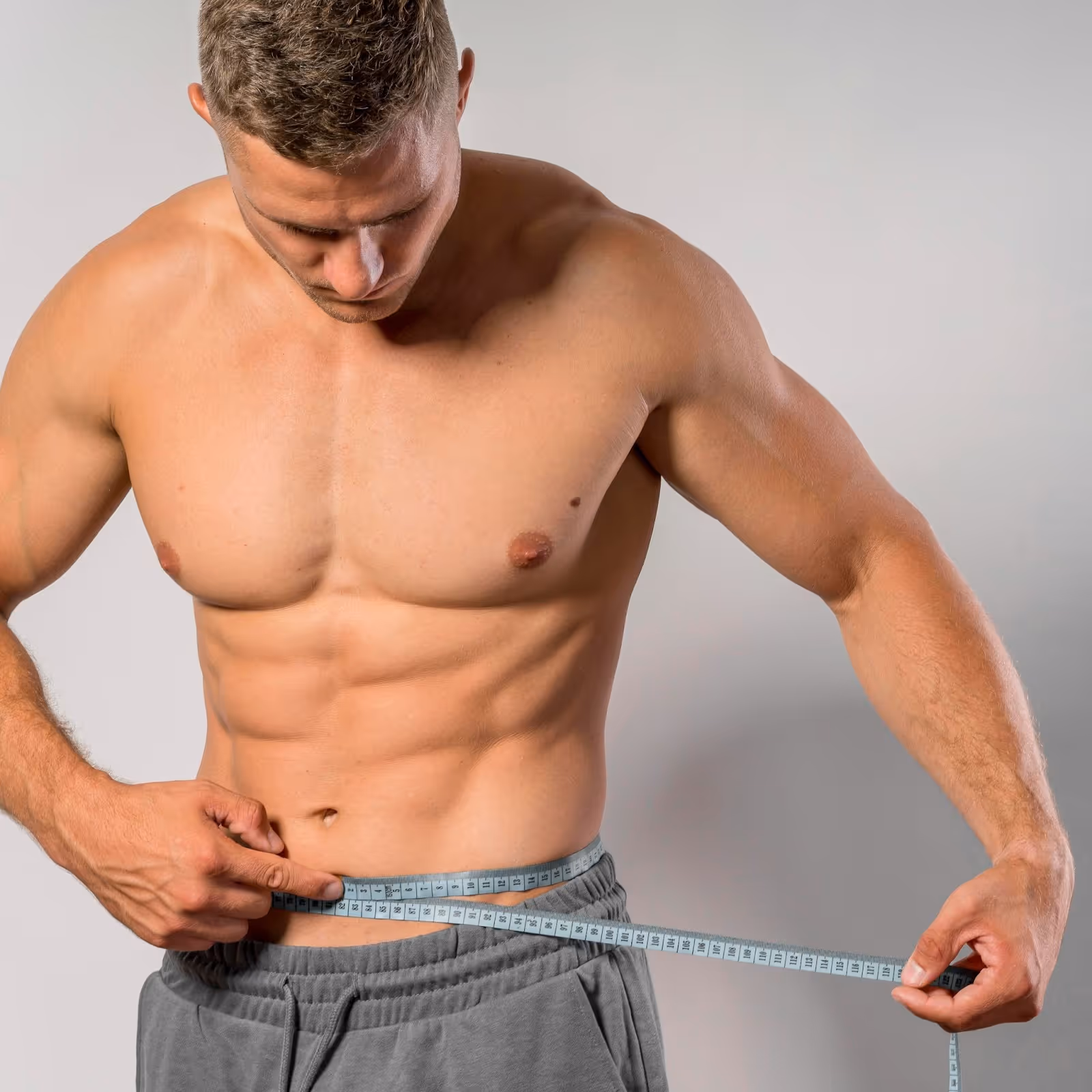 Fit athletic man measuring waist with measuring tape around defined abdominal area, showing body composition improvements and weight loss