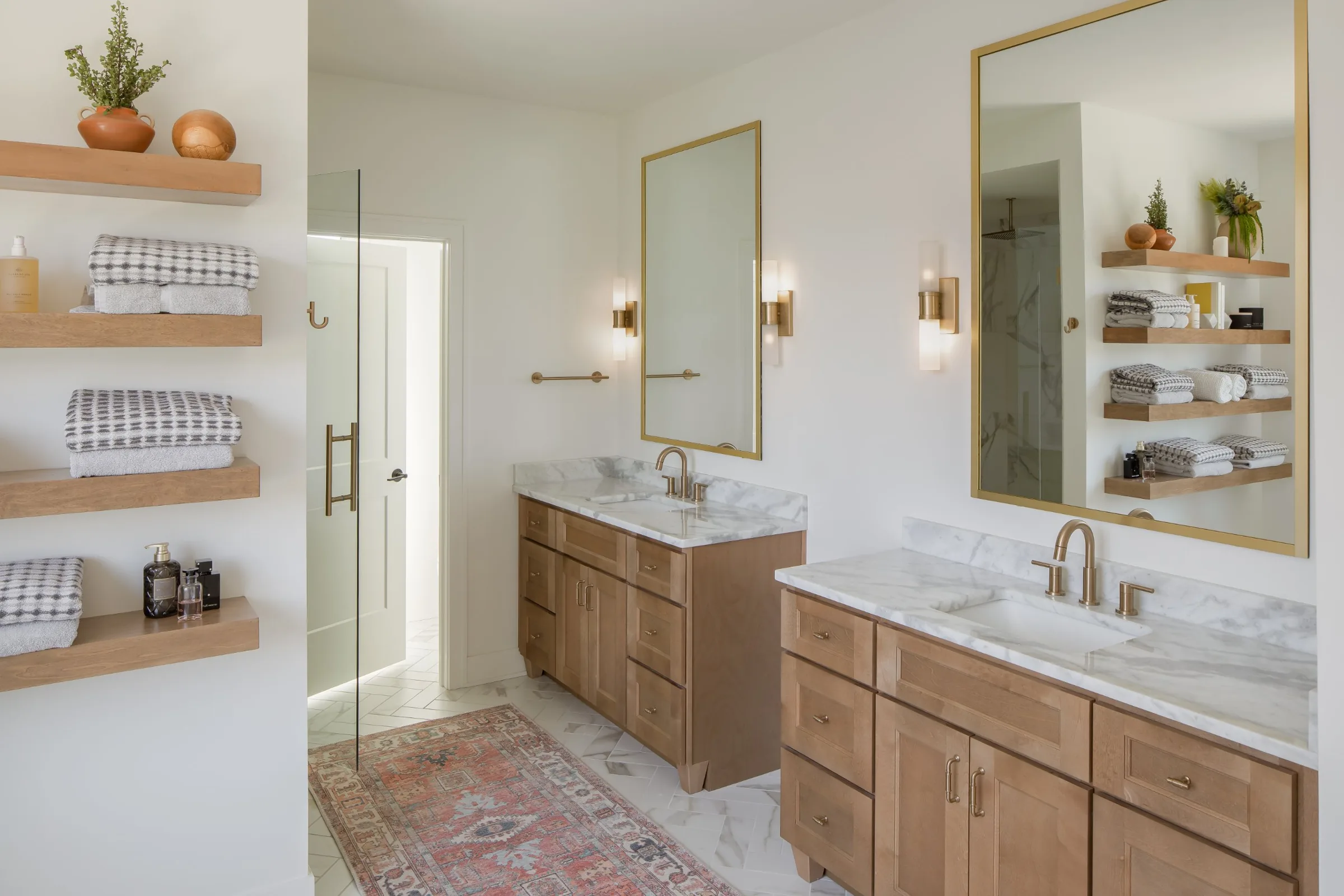 Bathroom with two wood vanities, marble-look tops, brass-framed mirrors, and floating wood shelves
