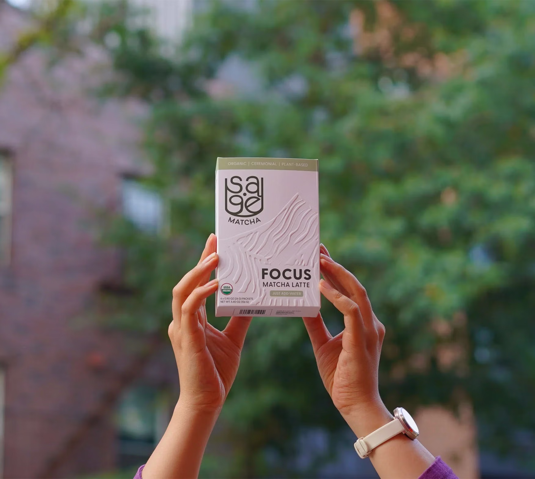 A pair of hands holding a box of Sage Matcha's Focus matcha blend up in the air with an out-of-focus brick building and tree in the background.