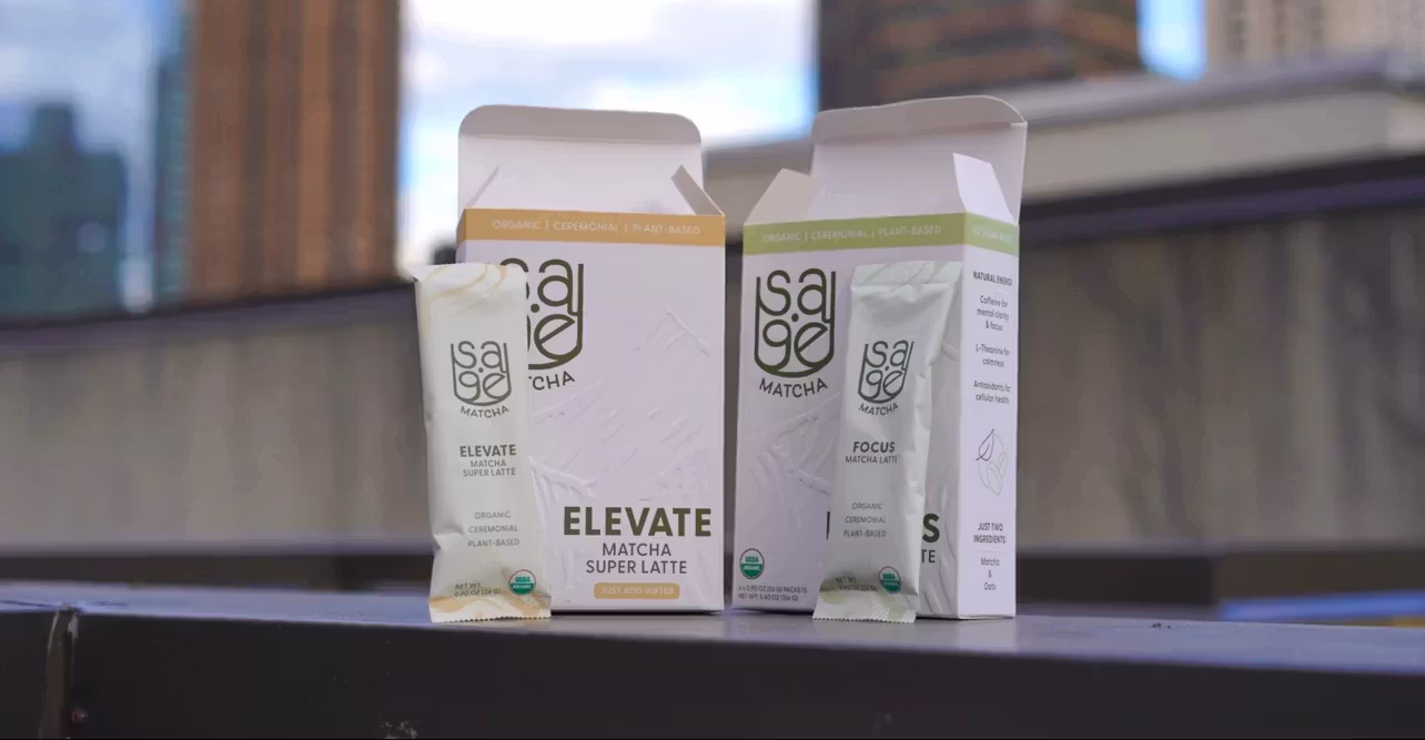 A staged product shot of both Sage Matcha blends' boxes with a sachet from each leaning against the front box panel on a ledge with an out-of-focus city in the background.