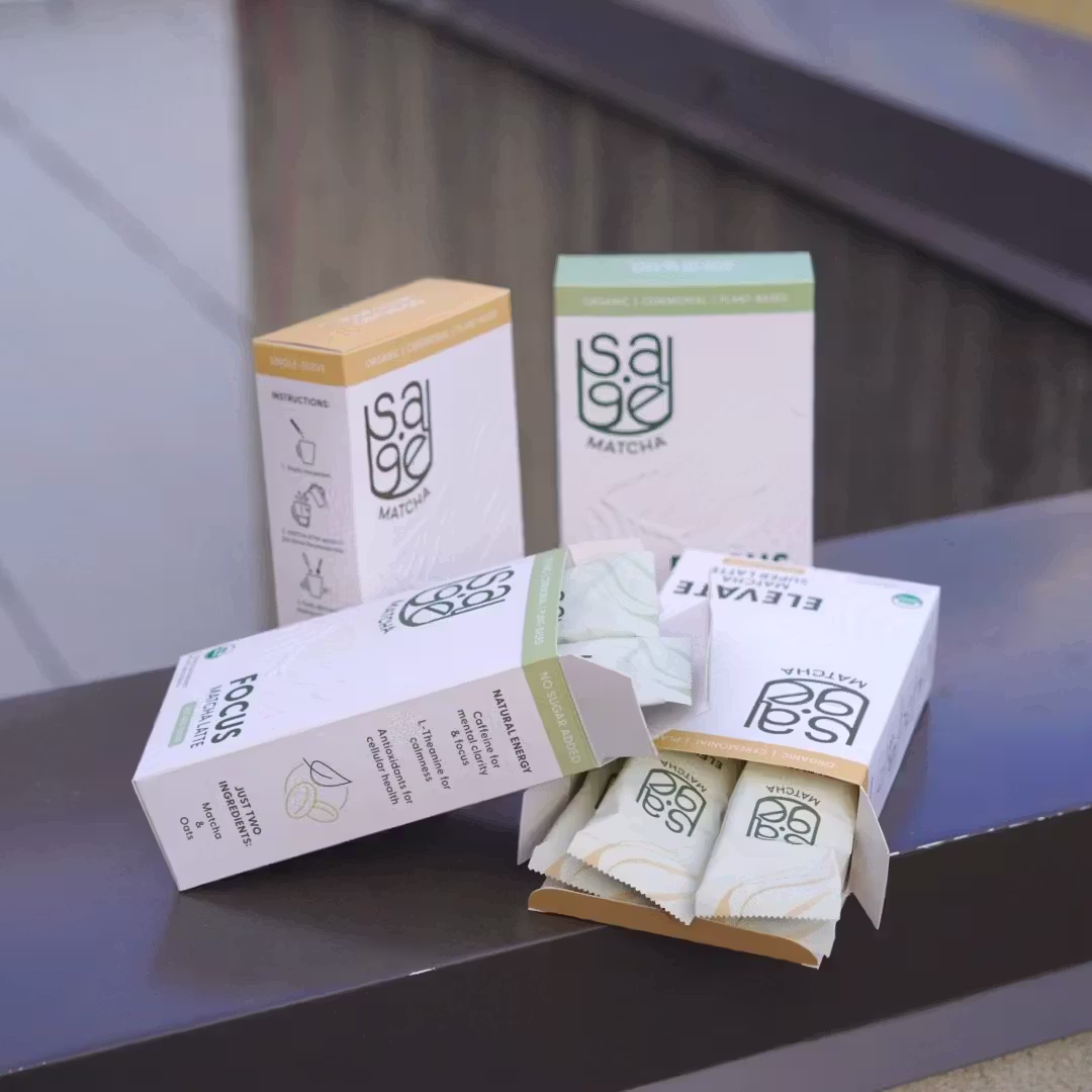 A staged product shot of both Sage Matcha blends' boxes on a metal ledge. Two boxes are standing upright in the back, while the other two boxes are open with the front box panel facing up, showing the sachets inside.