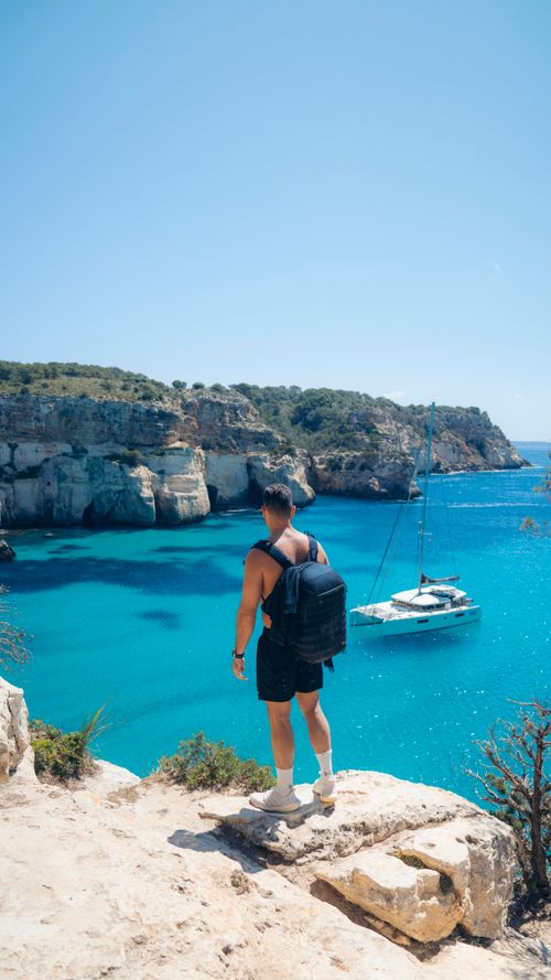 Man with backpack standing on a rocky cliff overlooking a turquoise sea with a sailboat near the shore.