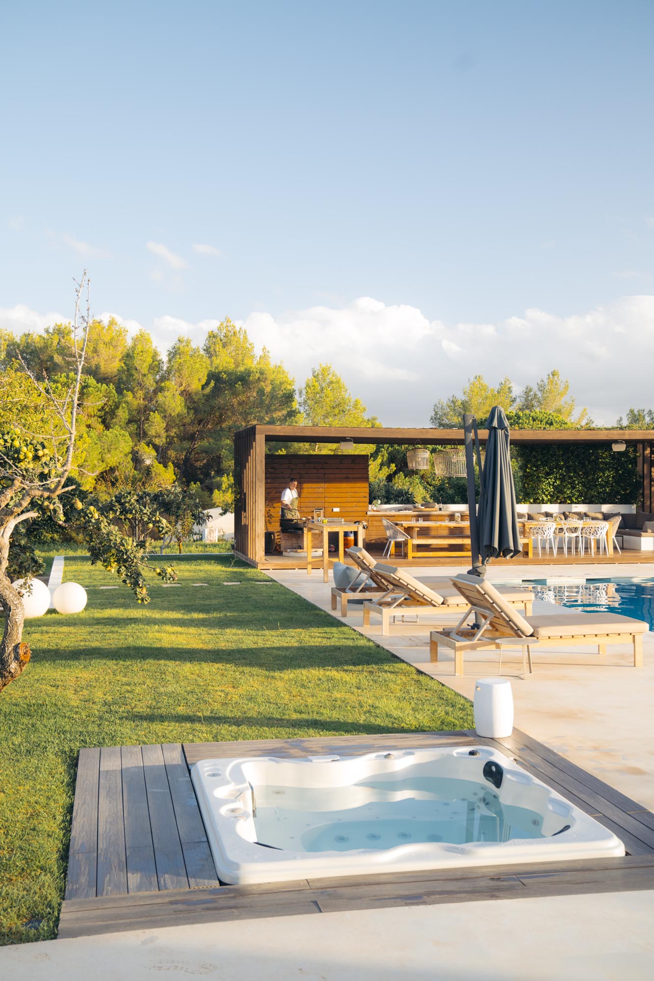 Outdoor scene with a sunlit Jacuzzi, lawn, poolside lounge chairs, and a man standing under a wooden pergola with dining and seating areas.