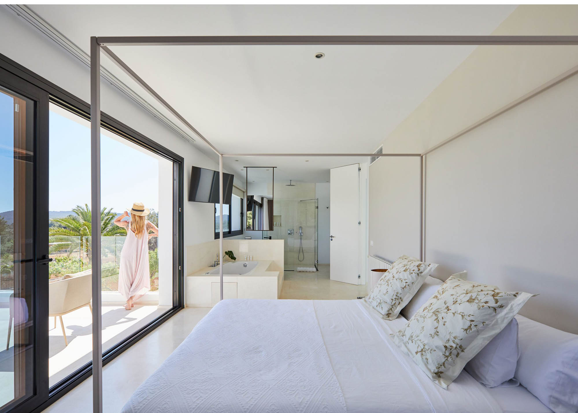 Bright modern bedroom with a four-poster bed, white and floral pillows, large sliding glass doors opening to a balcony where a woman in a hat stands, and an integrated bathroom with bathtub and shower.