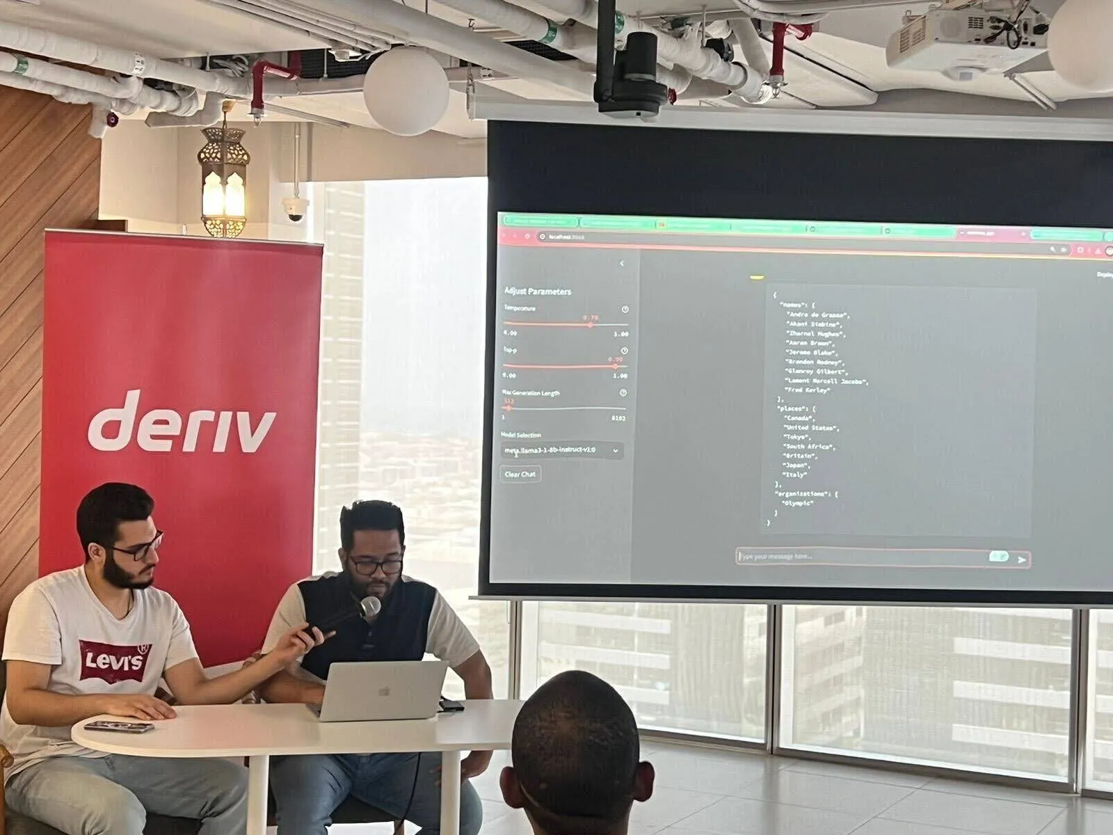 Deriv host holds a microphone while the AWS speaker presents on a laptop during an AWS community event.