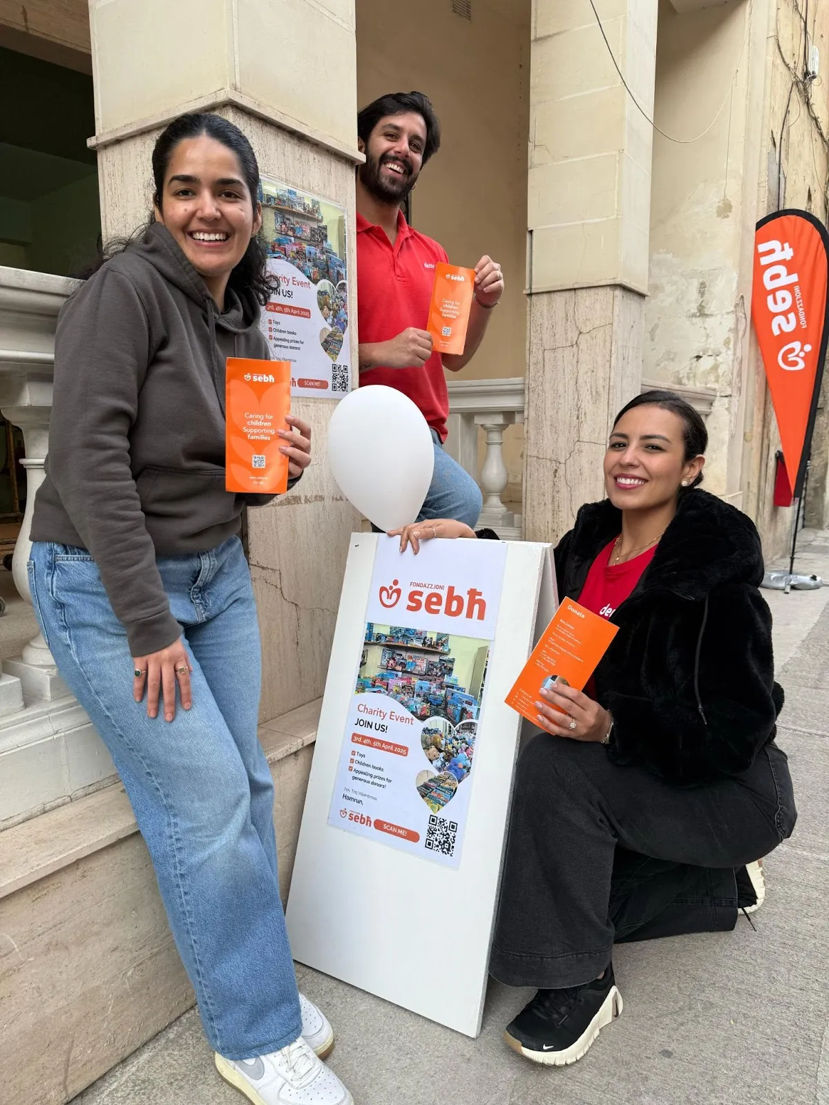 3 smiling volunteers hold flyers and a balloon while promoting a Deriv and Fondazzjoni Sebħ event outside a stone building.