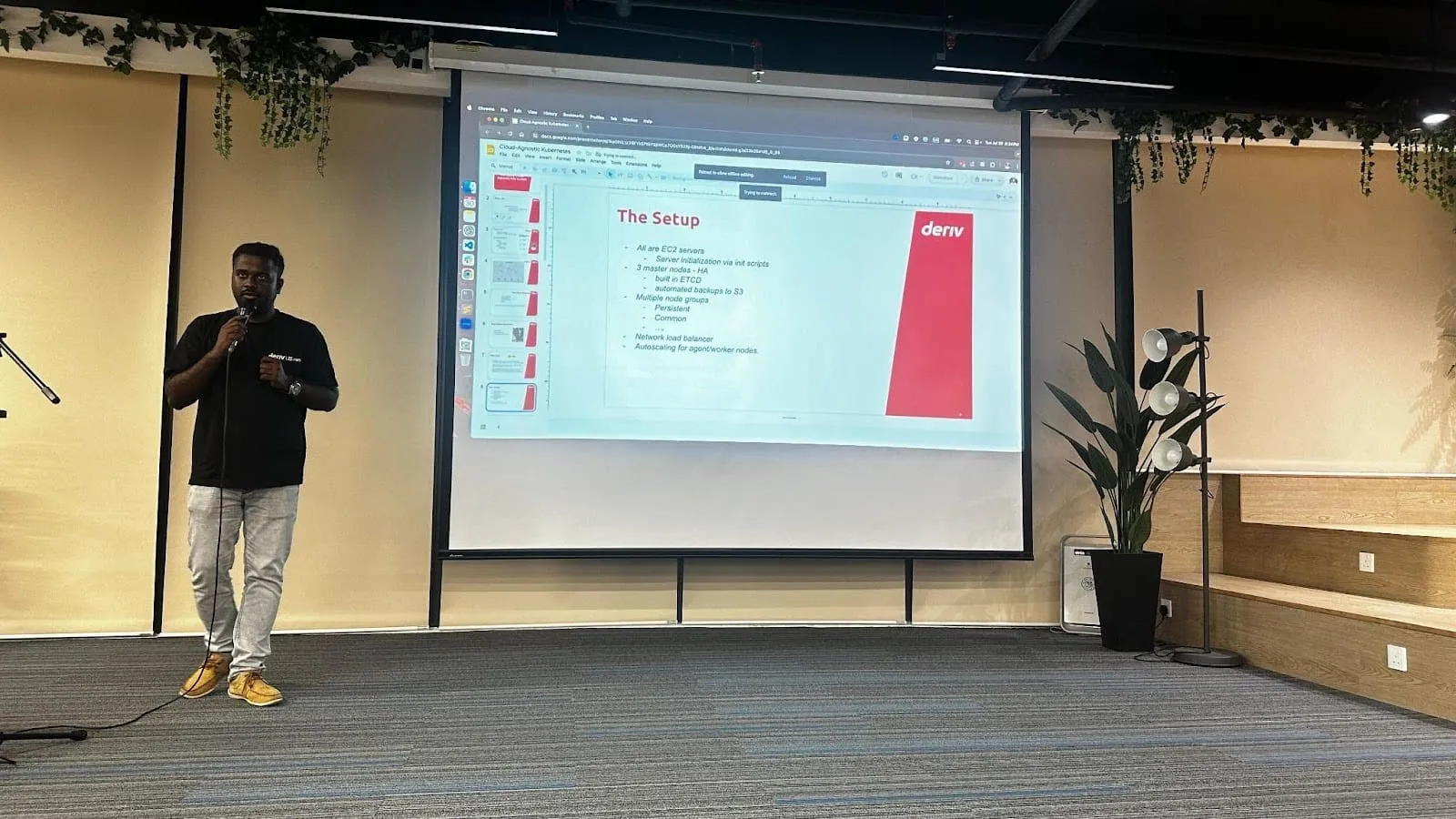 جوثيماني رادهاكريشنان، مهندس DevOps الرئيسي في Deriv، يتحدث على المنصة وخلفه شريحة عرض.