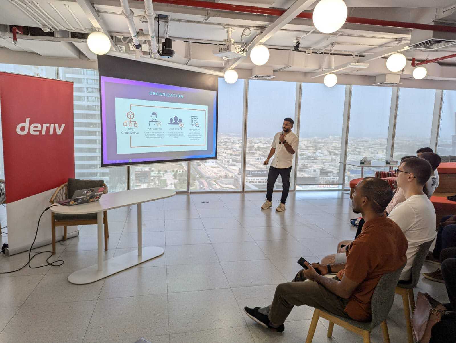 المُقدّم يتحدث عن AWS Organizations في حدث للتواصل، مع جمهور ولوحة Deriv ظاهرة.