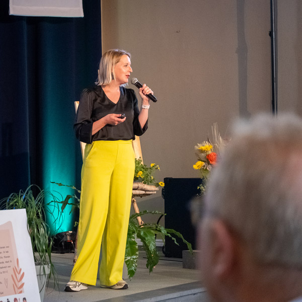 Een spreker in felgele broek en zwarte blouse richt zich tot het publiek tijdens de academische zitting voor 75 jaar Ons Kinderhuis.
