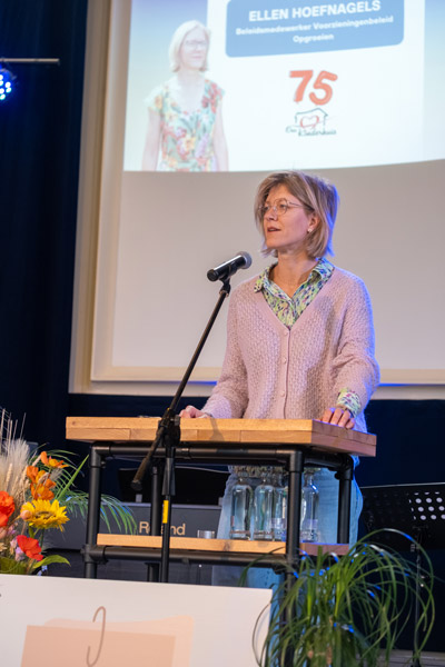 Ellen Hoefnagels spreekt het publiek toe vanaf het spreekgestoelte, met haar naam en functie geprojecteerd achter haar tijdens de viering.