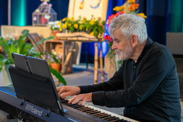 Een pianist speelt live muziek op een Roland-toestel tijdens het jubileumevenement van Ons Kinderhuis, omringd door bloemen.