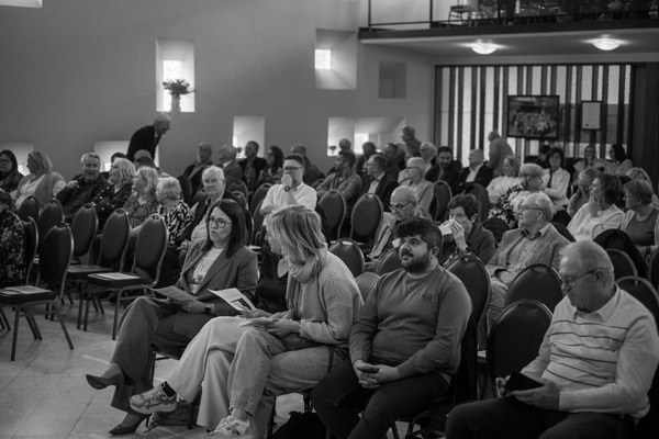 Een zwart-witfoto van een gevulde zaal met aandachtige toeschouwers tijdens de academische zitting voor het 75-jarig jubileum van Ons Kinderhuis. Mensen lezen het programmaboekje en wachten op de start van het evenement.