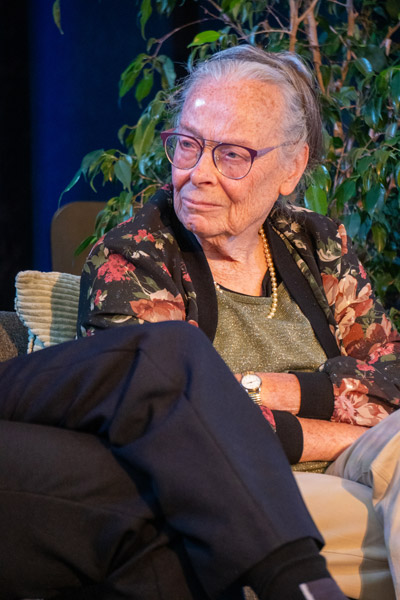 Een oudere vrouw met bril en bloemenvest luistert aandachtig tijdens het academisch panel ter ere van 75 jaar Ons Kinderhuis.