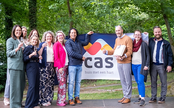 Een groep collega’s en betrokkenen poseert samen voor een CAS-banner tijdens de viering van de nieuwe naam van Ons Kinderhuis.