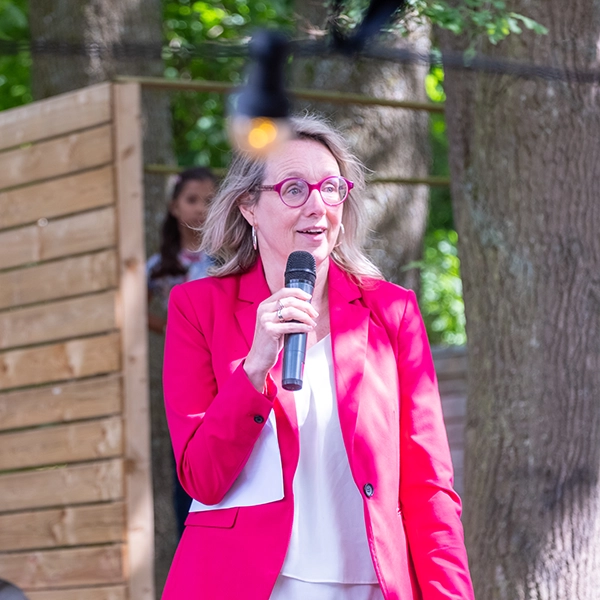Een vrouw in felroze blazer spreekt het publiek toe met een microfoon tijdens de naamsonthulling van CAS, buiten tussen de bomen.
