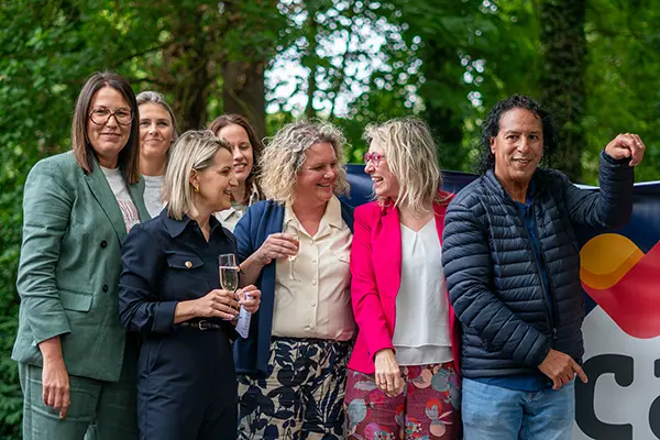 Groepsfoto van medewerkers en betrokkenen van Ons Kinderhuis, lachend bij een CAS-banner tijdens de naamsonthulling in een groene buitenomgeving.