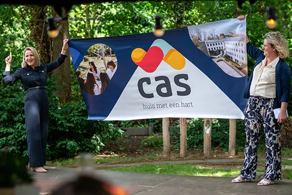 Twee vrouwen houden een grote banner omhoog met het nieuwe logo en de naam “CAS – huis met een hart” tijdens de officiële onthulling.