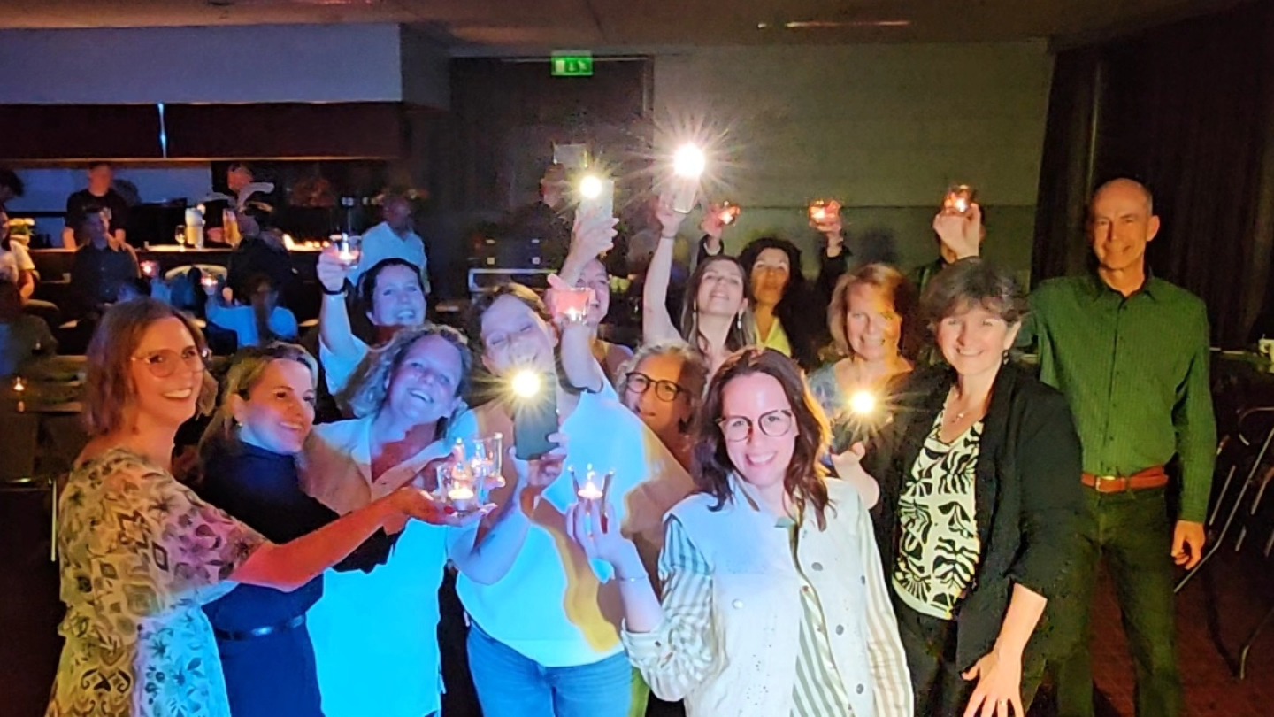 Een groep medewerkers houdt kaarsjes en telefoonlampjes omhoog in een donkere zaal, lachend en samen het jubileum vierend.