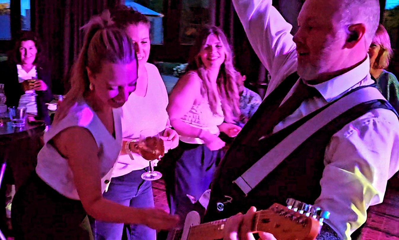 Een gitarist speelt in het publiek terwijl feestvierders met een drankje in de hand enthousiast meebewegen op de muziek.