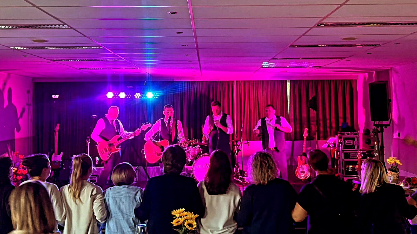 Een liveband treedt op op een verlichte paarse podium in een zaal, terwijl een enthousiast publiek danst en meeklapt.

