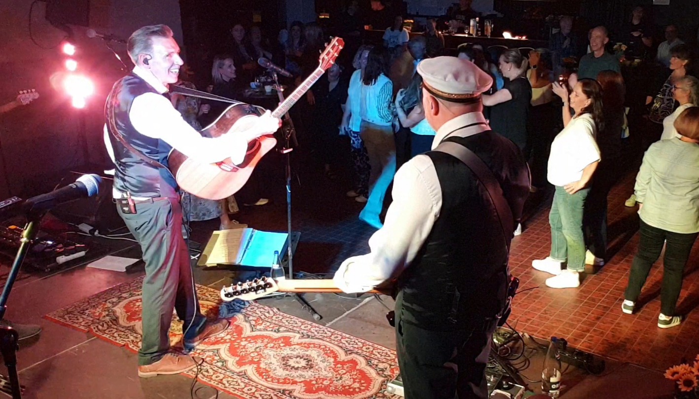 Twee bandleden met gitaren op het podium kijken glimlachend naar een dansende menigte tijdens het personeelsfeest.
