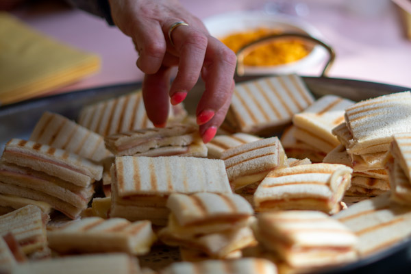 Een hand pakt een gegrilde sandwich van een schaal met hapjes, klaar voor de gasten van het tuinfeest.