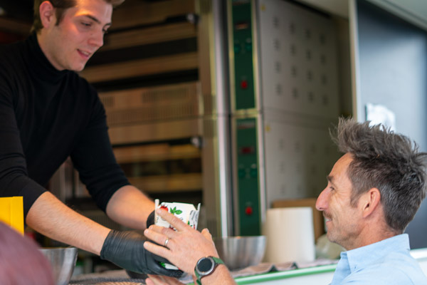 Een medewerker van een foodtruck reikt met een glimlach een vers gerecht aan een bezoeker tijdens het tuinfeest.