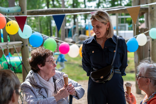 Een jonge vrouw in donkerblauw praat met twee oudere dames die een ijsje eten, tegen een kleurrijke achtergrond met slingers en ballonnen.