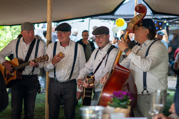 Een vierkoppige folkgroep speelt live muziek onder een feesttent, gekleed in witte hemden, bretellen en petten.