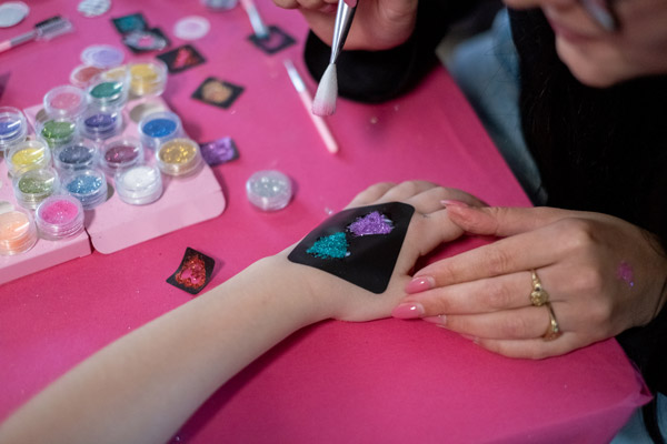 Een glittertattoo wordt aangebracht op de hand van een kind, met potjes kleurige glitters en sjablonen op een roze tafel.