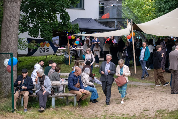 Bezoekers van het tuinfeest zitten op bankjes en wandelen onder tentzeilen, met het CAS-logo zichtbaar op de achtergrond.