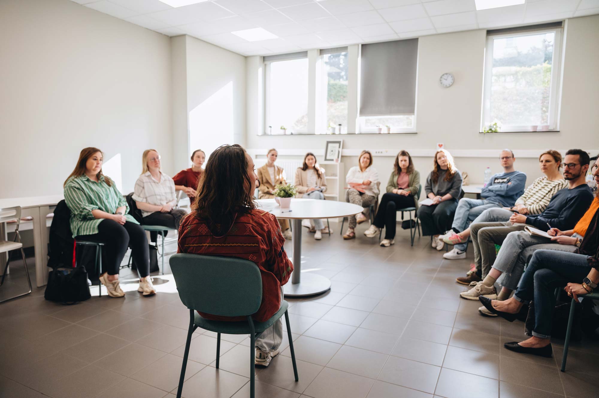 Een groep volwassenen zit in een kring in een lichte, moderne vergaderruimte tijdens een groepsgesprek of teamvergadering. De sfeer oogt betrokken en open. Eén persoon, met de rug naar de camera, spreekt de rest toe. Grote ramen zorgen voor natuurlijk licht