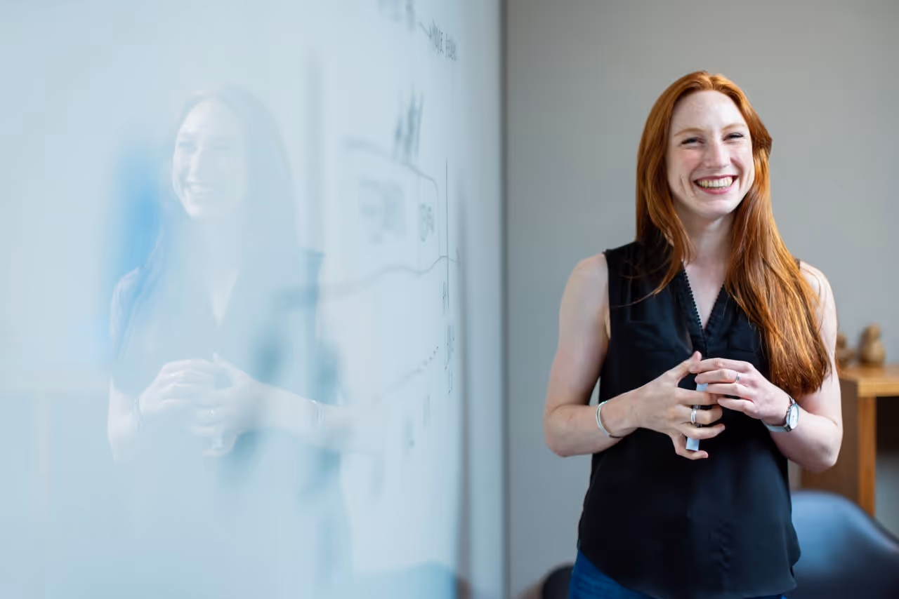 Kvinde smiler foran en tavle, der viser hendes engagement og positive attitude i et læringsmiljø.