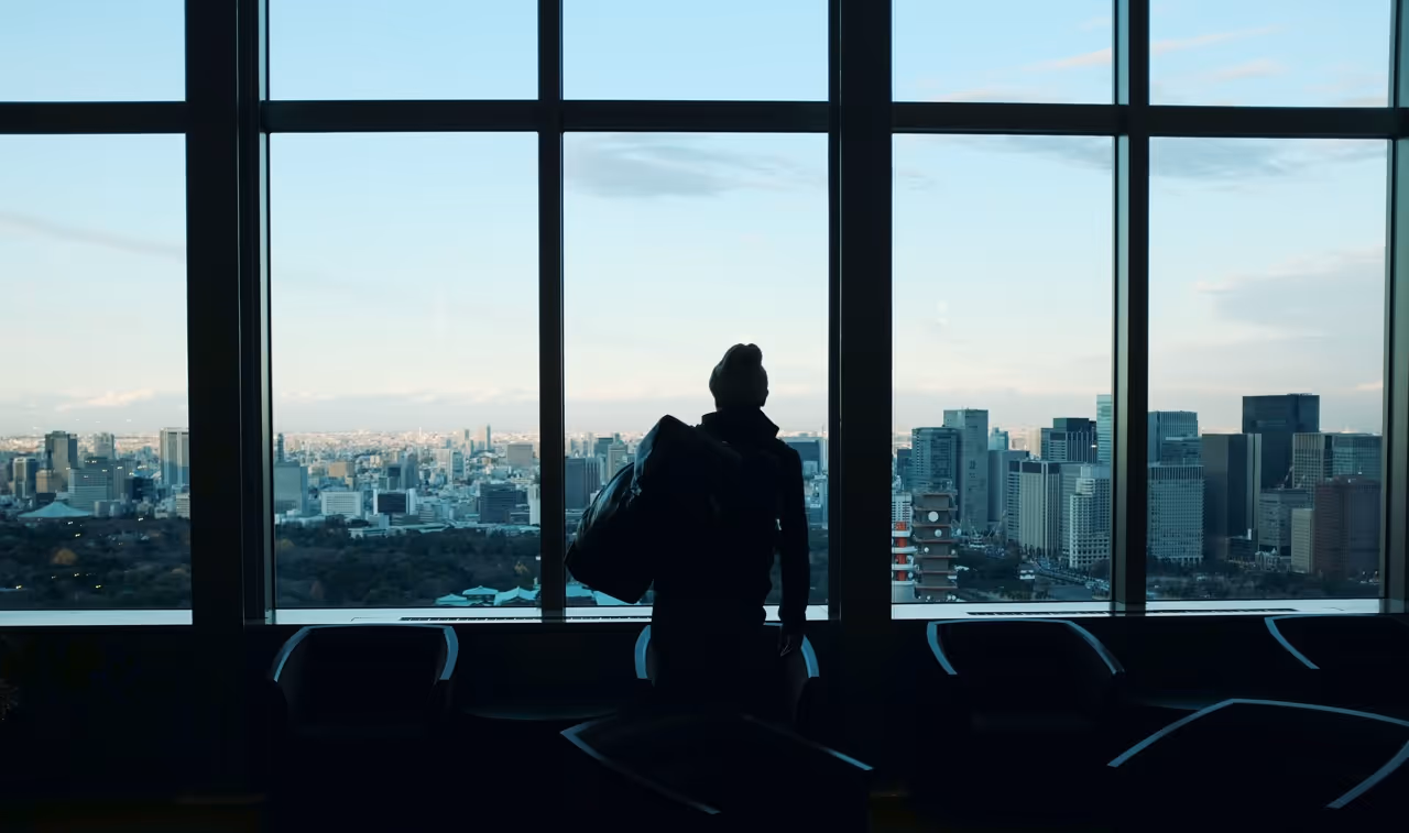 Person står foran et vindue og ser ud på en skyline af bygninger, omgivet af naturligt lys og en rolig atmosfære.