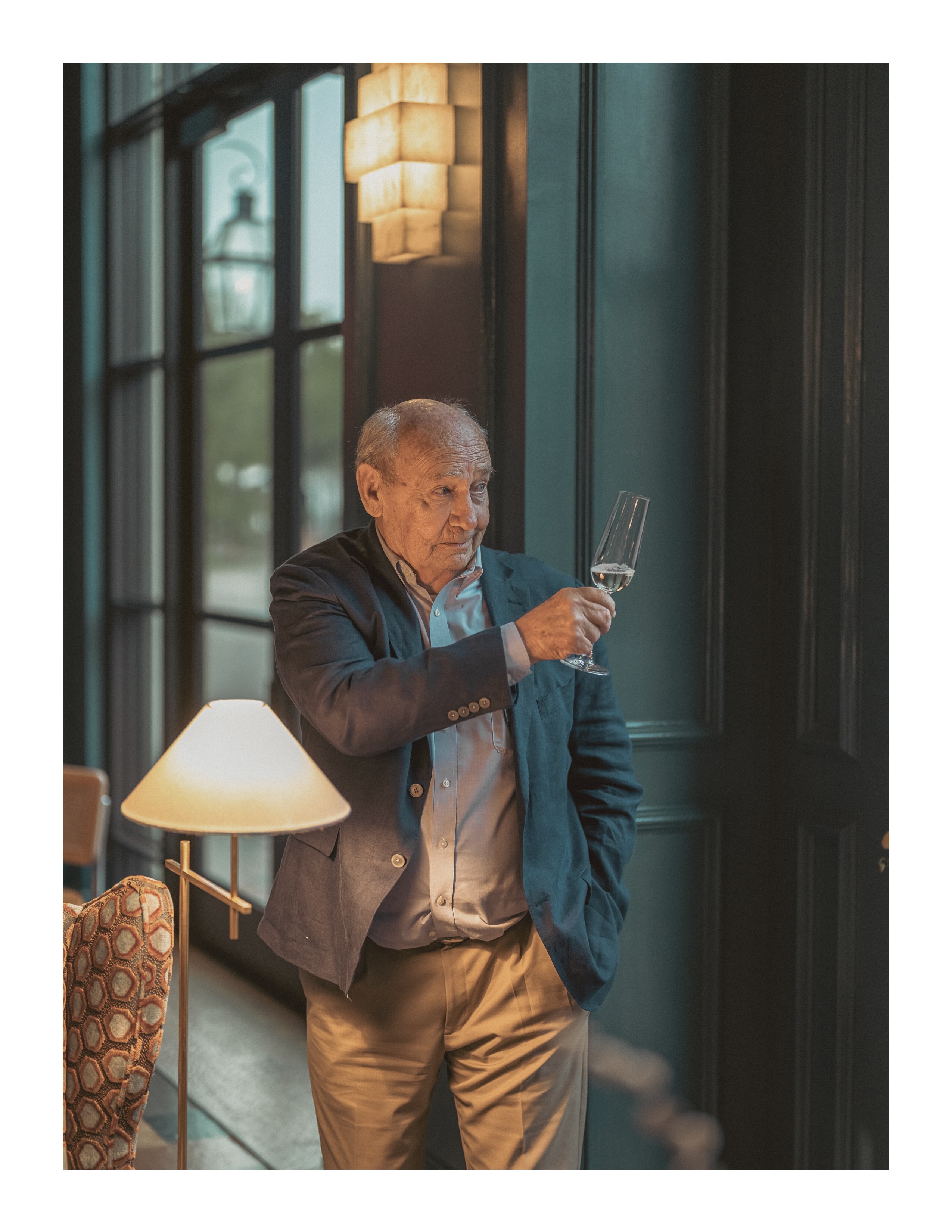 Elderly man in a navy blazer holding a glass of champagne inside a warmly lit hotel bar with green paneling