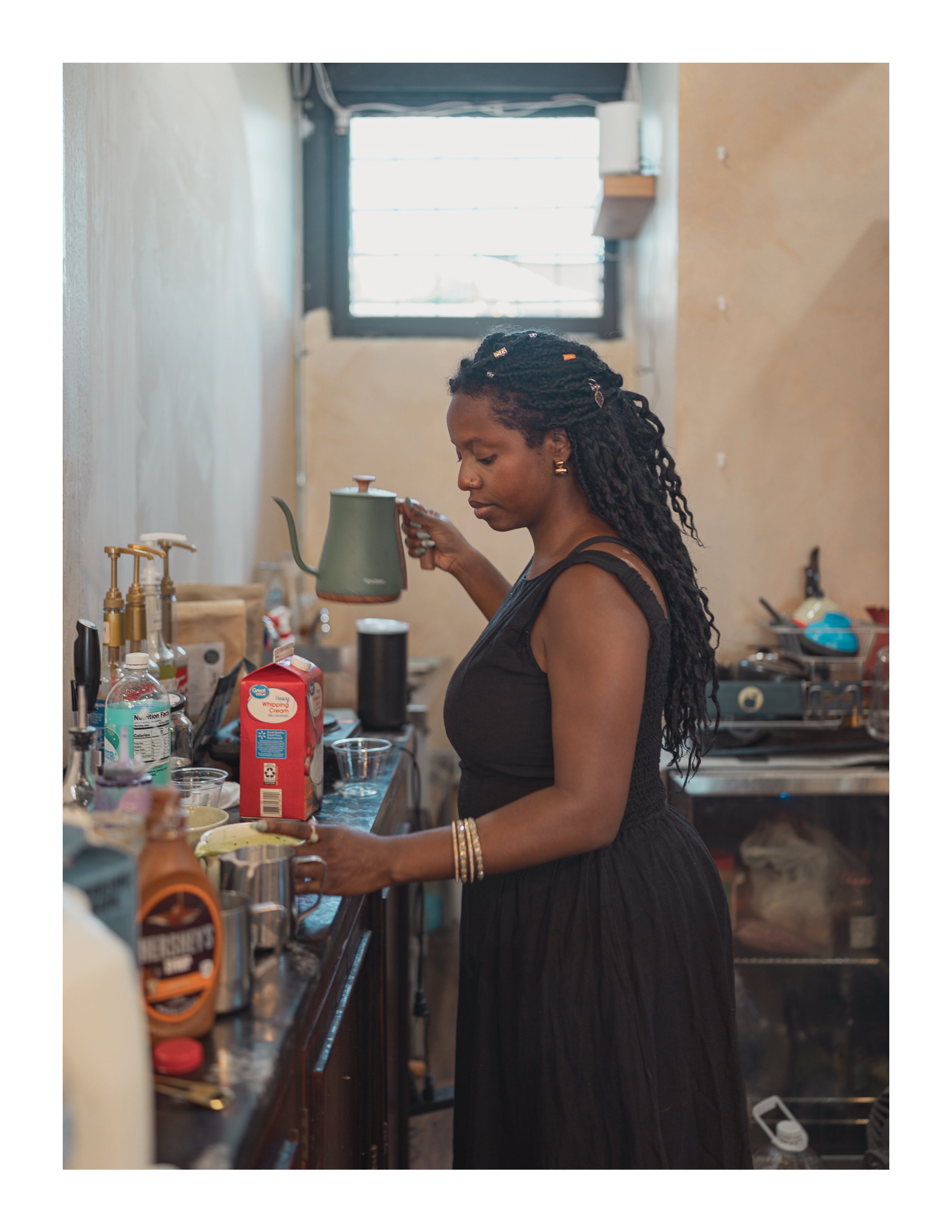 A woman in a black dress pours hot water from a kettle in a cozy cafe, surrounded by ingredients and morning light.