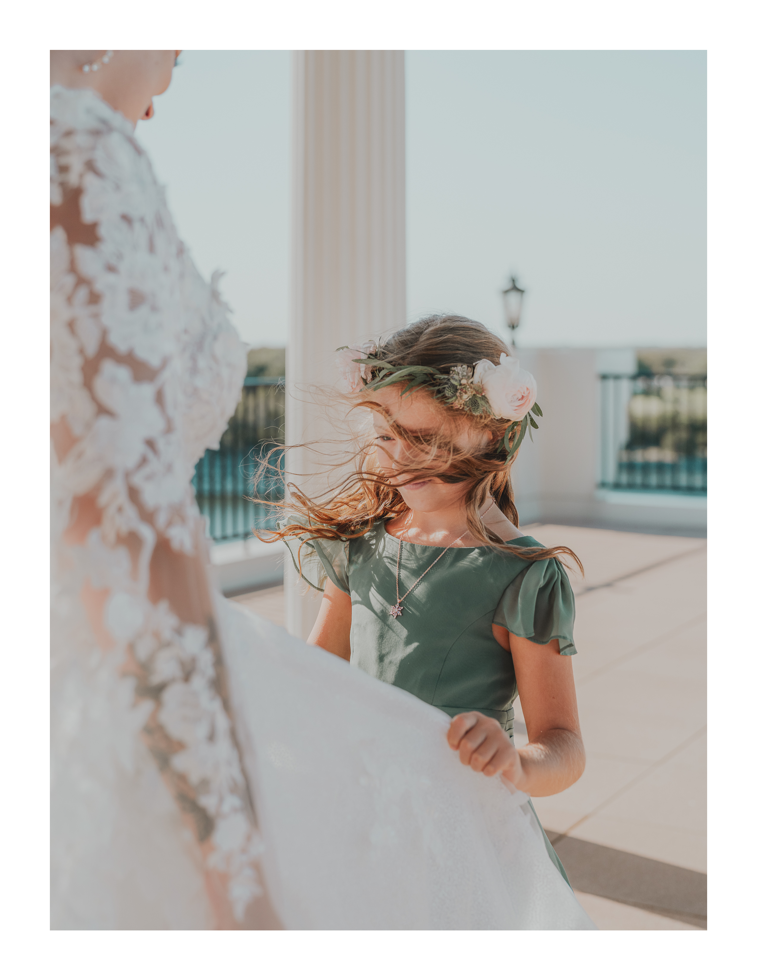 A young flower girl holds the bride’s dress as wind blows her hair across her face during a sunlit outdoor ceremony.