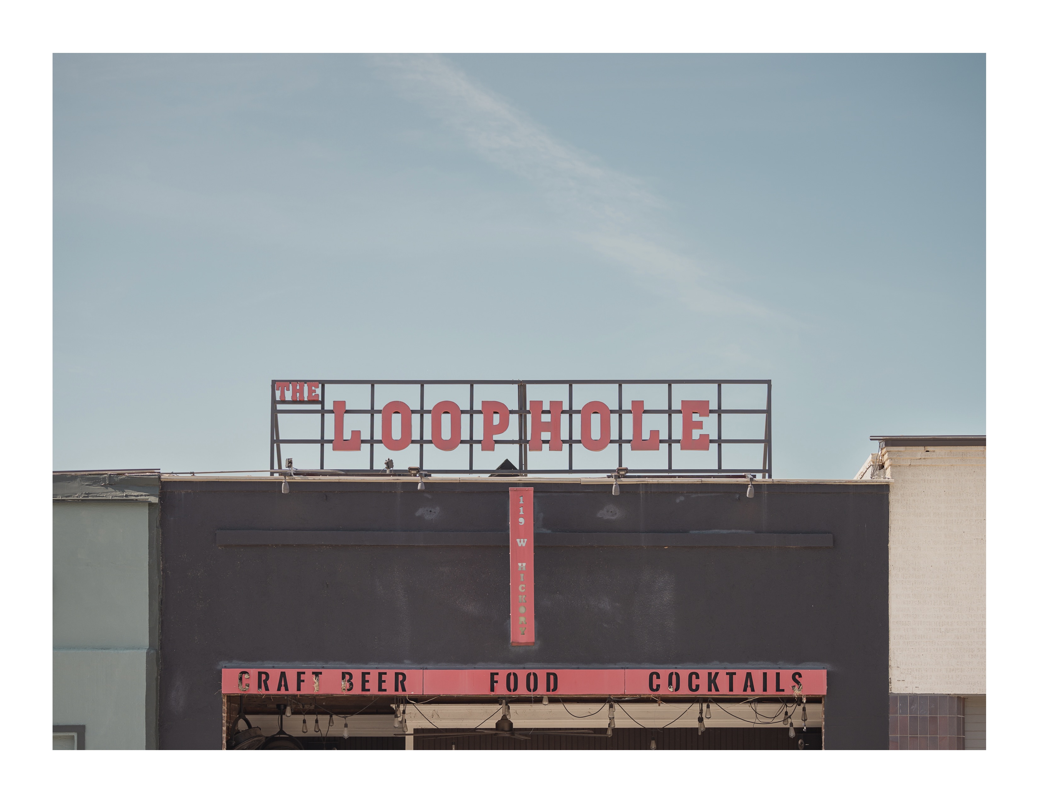 Red sign reading “The Loophole” sits atop a dark building with a retro facade, photographed under soft morning light.