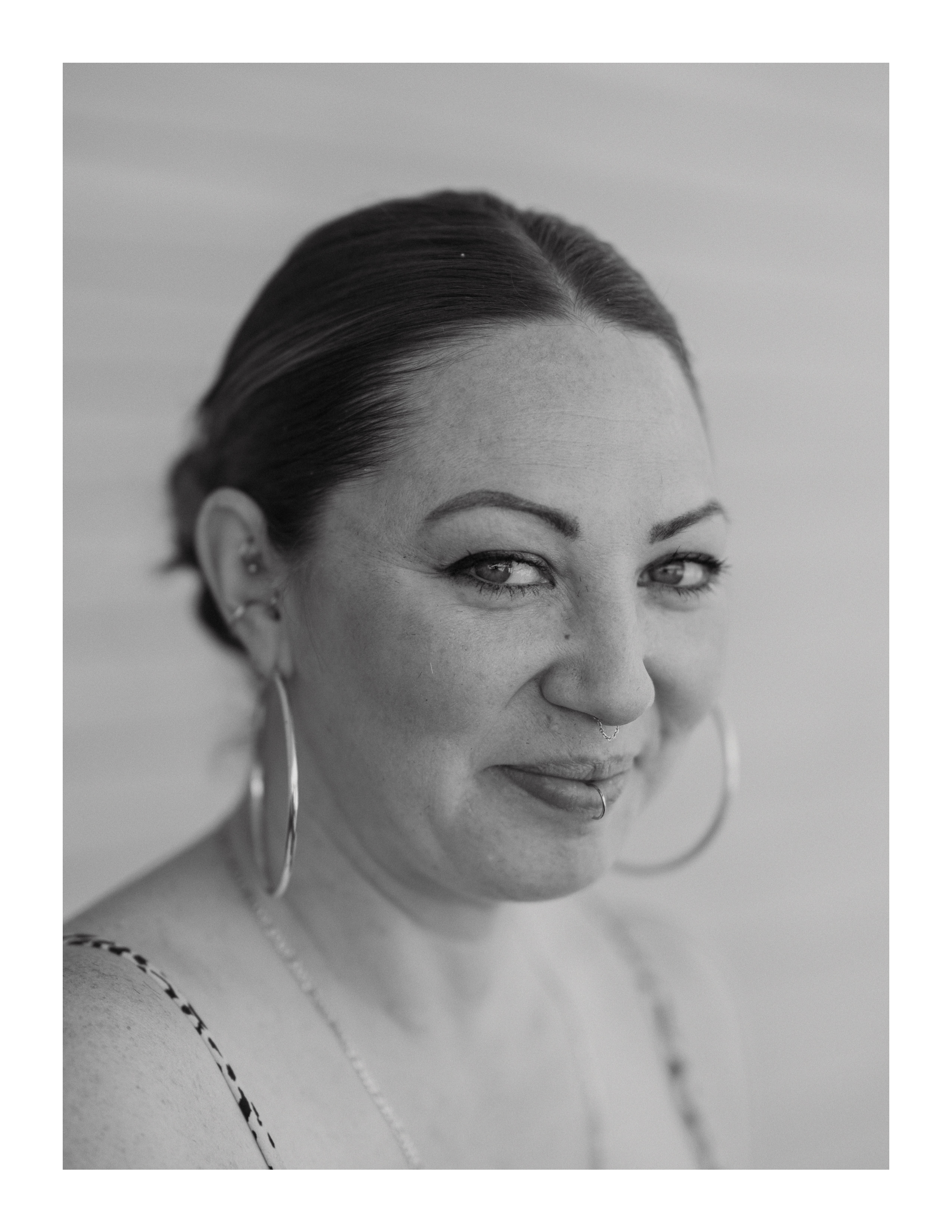 Close-up portrait of a woman smiling softly, wearing hoop earrings and a lip ring, captured in black and white.