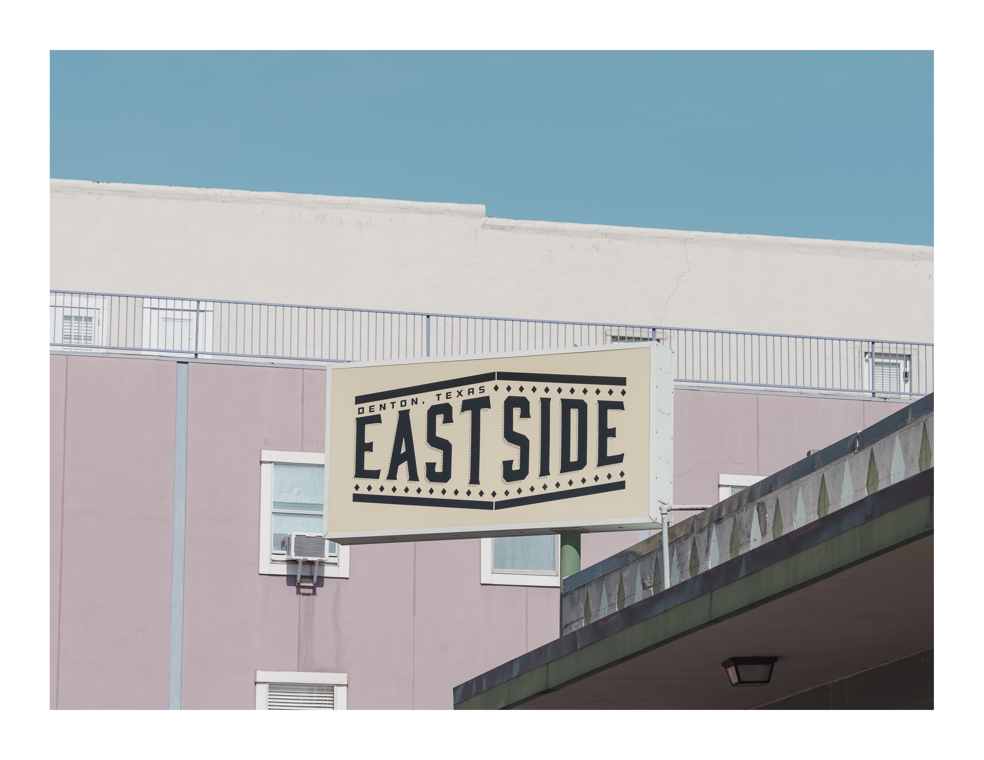 A cream and black “East Side” sign hangs against a pastel building facade in Denton, Texas, with a clear blue sky above.