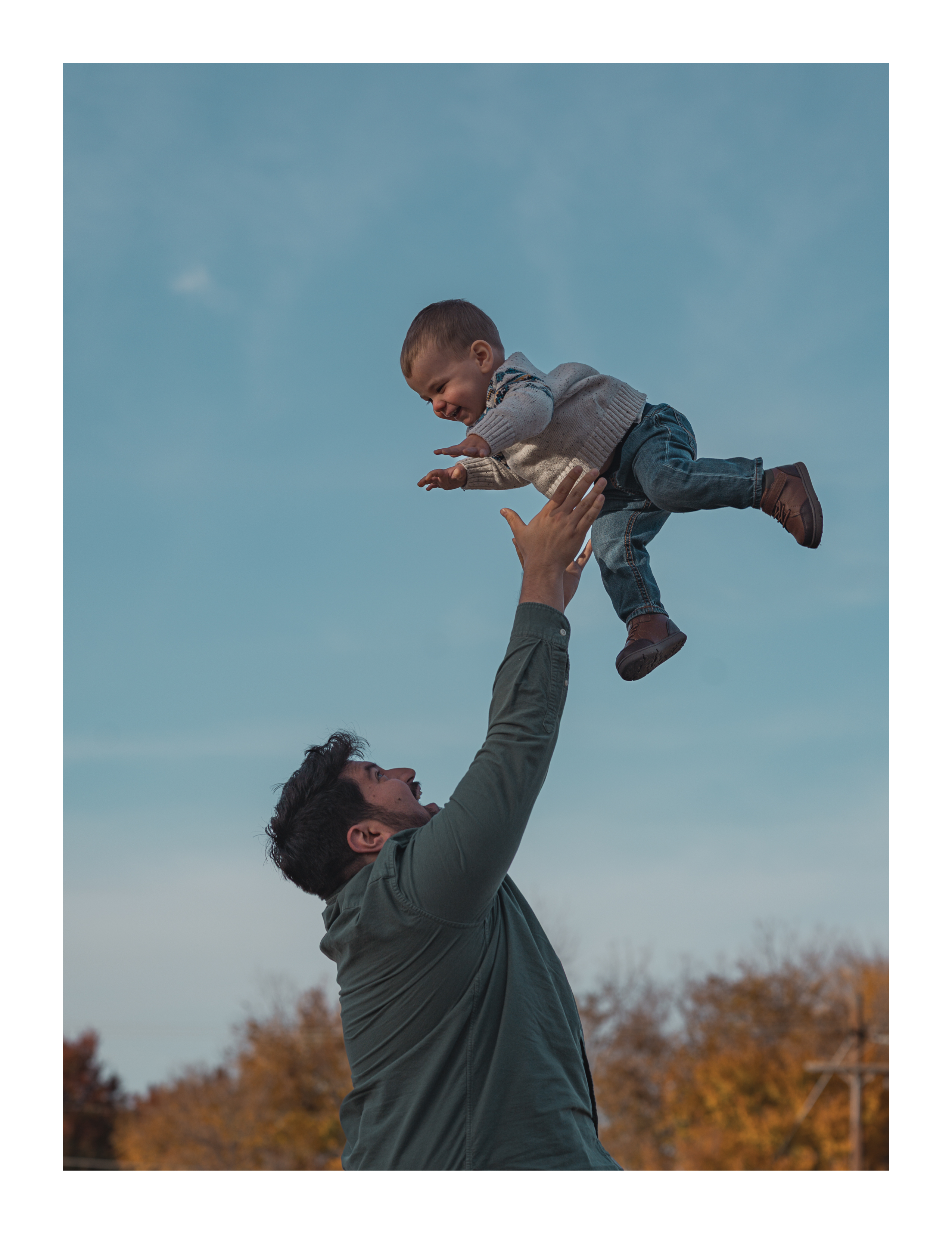 Father tossing son playfully in the air under open sky with fall foliage in background